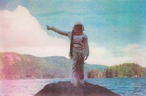 Image of a person in an old astronaut suit standing on a hill in front of a body of water surrounded by trees. The astronaut is pointing to the left.