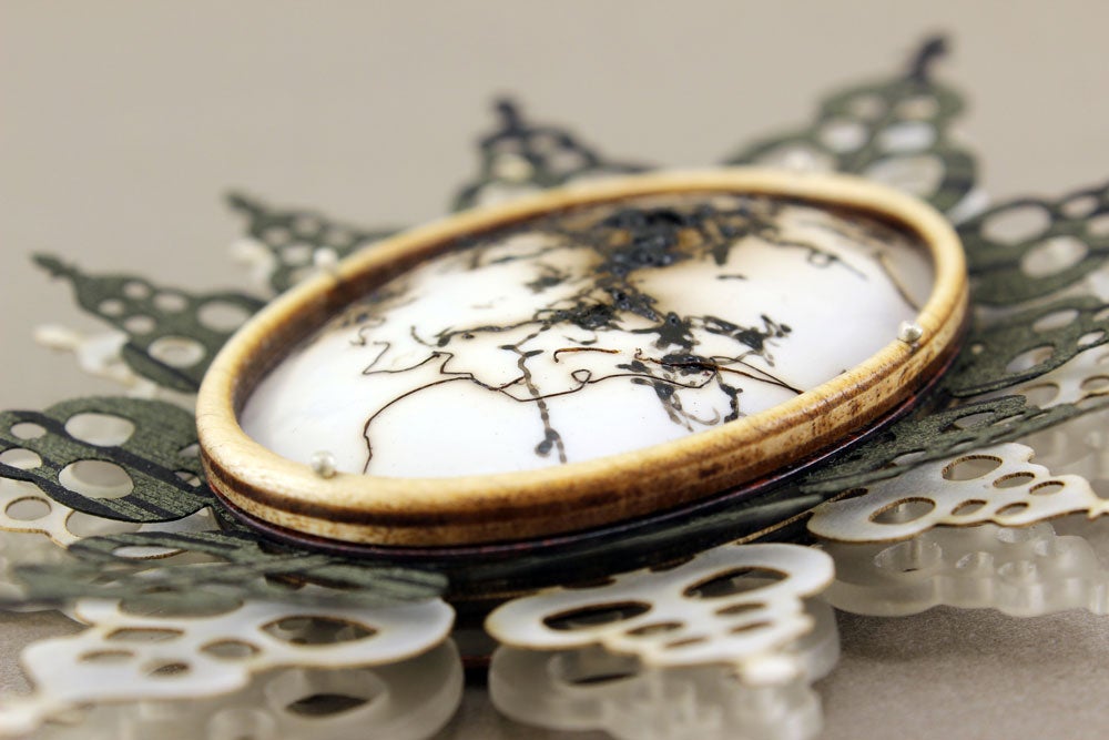 a white circle with what appears to be horse hair burned into it encased with a gold frame and geometric silver metal petals